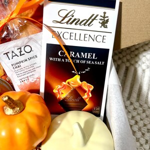 May include: A gift basket with a variety of fall-themed items, including a pumpkin spice chai tea bag, a bar of Lindt Excellence Caramel with a touch of sea salt chocolate, and a small ceramic pumpkin.