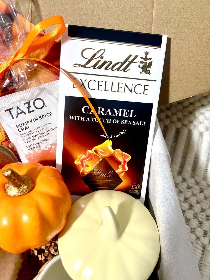 May include: A gift basket with a variety of fall-themed items, including a pumpkin spice chai tea bag, a bar of Lindt Excellence Caramel with a touch of sea salt chocolate, and a small ceramic pumpkin.