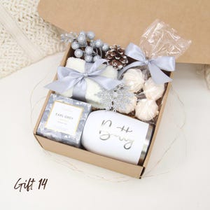 Puede incluir: Una caja de regalo llena de una taza blanca con la palabra "Betty", t&eacute;, una toalla peque&ntilde;a y dulces, todo atado con cintas plateadas. Acentos plateados decorativos y una pi&ntilde;a completan el arreglo. La caja est&aacute; etiquetada como "Gift 14".