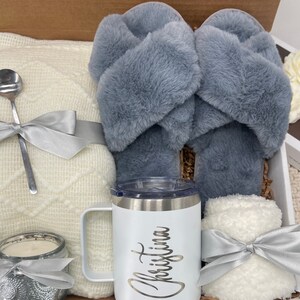 Op de afbeelding: Een cadeaudoos gevuld met knusse items, waaronder een wit deken, een paar grijze fluffy pantoffels, een witte mok met de naam "Christina" erop, een witte kaars en een witte fluffy deken.