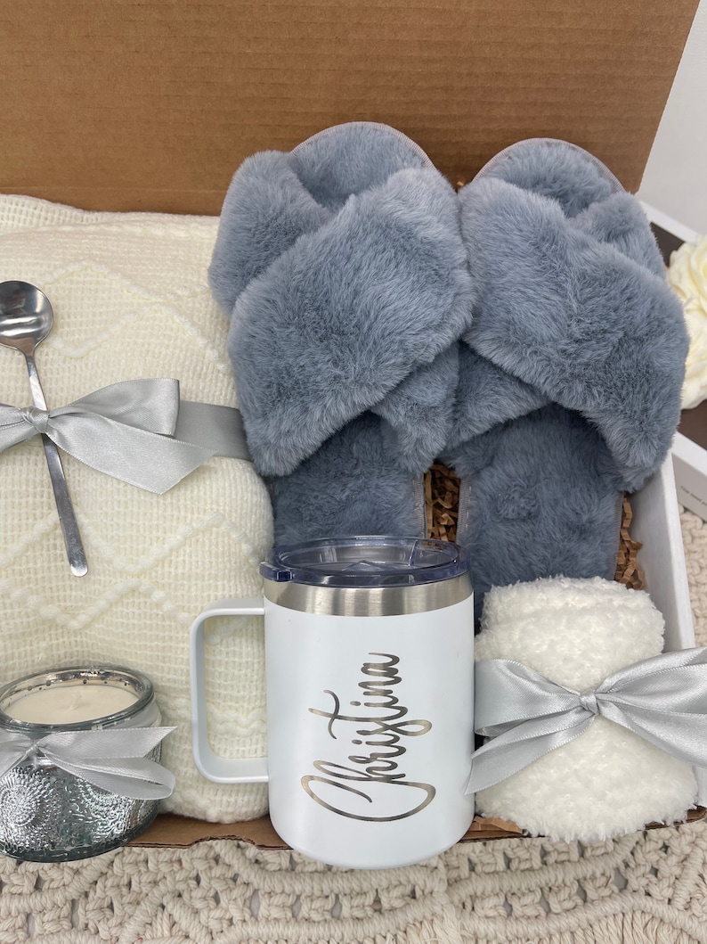 Op de afbeelding: Een cadeaudoos gevuld met knusse items, waaronder een wit deken, een paar grijze fluffy pantoffels, een witte mok met de naam "Christina" erop, een witte kaars en een witte fluffy deken.