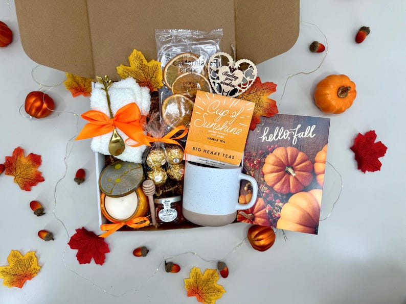 May include: A gift box filled with fall-themed items. Includes a mug, tea, a candle, a small towel with a spoon, dried orange slices, chocolates, honey, and a card that says "hello, fall". Decorative leaves and pumpkins surround the box.