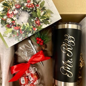 Pu&ograve; includere: Una scatola regalo piena di un bicchiere nero e argento con il testo "Mr. & Mrs." in lettere dorate, un fiocco rosso, un biglietto con una ghirlanda di agrifoglio innevata e una piccola borsa di cioccolatini rossi.