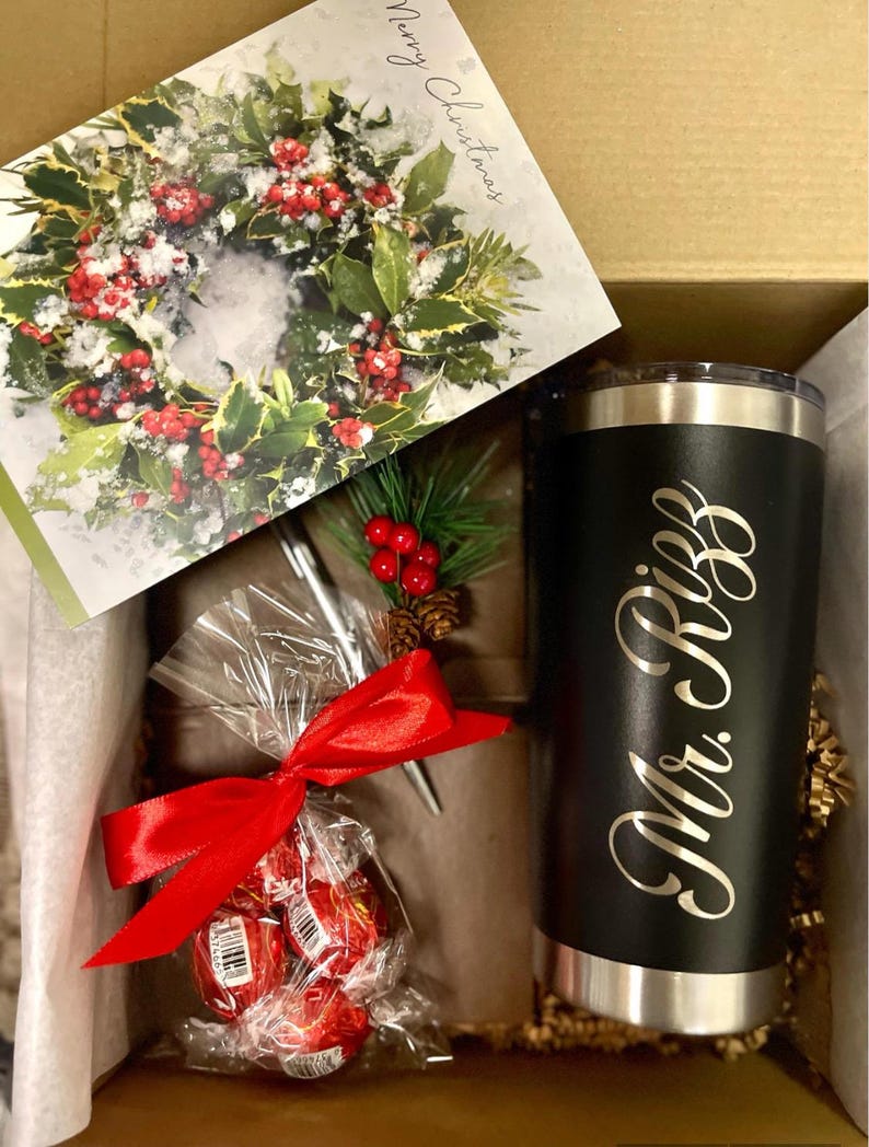 Pu&ograve; includere: Una scatola regalo piena di un bicchiere nero e argento con il testo "Mr. & Mrs." in lettere dorate, un fiocco rosso, un biglietto con una ghirlanda di agrifoglio innevata e una piccola borsa di cioccolatini rossi.
