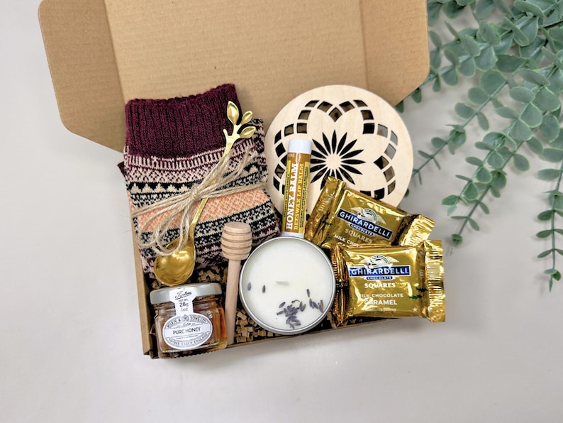 May include: A gift box filled with cozy items. Includes a pair of burgundy patterned socks, a gold-colored spoon, a small jar of honey, a wooden honey dipper, a candle, lip balm, a wooden coaster, and Ghirardelli chocolate squares.