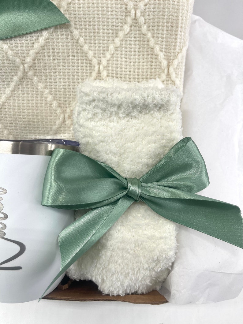 May include: A gift basket with a white fuzzy blanket, a white mug with a black design, and a green satin ribbon.
