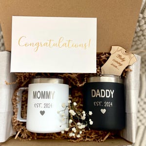 Puede incluir: Caja de regalo con una taza blanca y otra negra, una tarjeta con "Congratulations!" y una etiqueta de madera con "Baby coming soon...". La taza blanca dice "MOMMY EST. 2024" y la negra "DADDY EST. 2024".