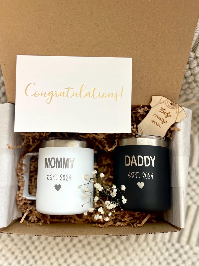 Puede incluir: Caja de regalo con una taza blanca y otra negra, una tarjeta con "Congratulations!" y una etiqueta de madera con "Baby coming soon...". La taza blanca dice "MOMMY EST. 2024" y la negra "DADDY EST. 2024".