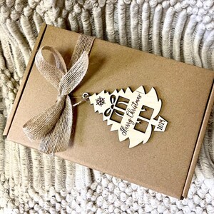 May include: A brown cardboard box with a rustic brown ribbon and a wooden Christmas tree ornament. The ornament is engraved with the text "Merry Christmas! 2024".