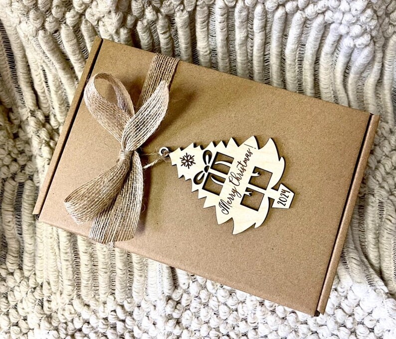 May include: A brown cardboard box with a rustic brown ribbon and a wooden Christmas tree ornament. The ornament is engraved with the text "Merry Christmas! 2024".