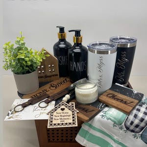 K&ouml;nnte beinhalten: Eine Holzkiste gef&uuml;llt mit Einweihungsgeschenken, darunter ein hausf&ouml;rmiges Holzschild, zwei schwarz-wei&szlig;e Handseifenflaschen, zwei personalisierte Becher, eine Kerze, ein Holzschl&uuml;sselanh&auml;nger, ein Holzhaus-Anh&auml;nger und ein gr&uuml;n-wei&szlig; gestreiftes Geschirrtuch.