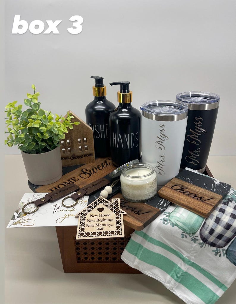 K&ouml;nnte beinhalten: Eine Holzkiste gef&uuml;llt mit Einweihungsgeschenken, darunter ein hausf&ouml;rmiges Holzschild, zwei schwarz-wei&szlig;e Handseifenflaschen, zwei personalisierte Becher, eine Kerze, ein Holzschl&uuml;sselanh&auml;nger, ein Holzhaus-Anh&auml;nger und ein gr&uuml;n-wei&szlig; gestreiftes Geschirrtuch.