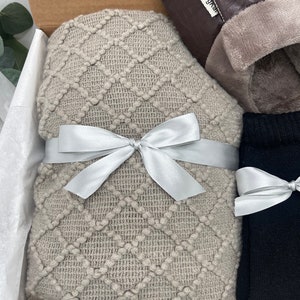 Pode incluir: Uma caixa de presente cheia de itens aconchegantes, incluindo uma manta de malha bege com uma fita prateada, um par de meias pretas e um par de pantufas marrons com uma etiqueta "JingKea".