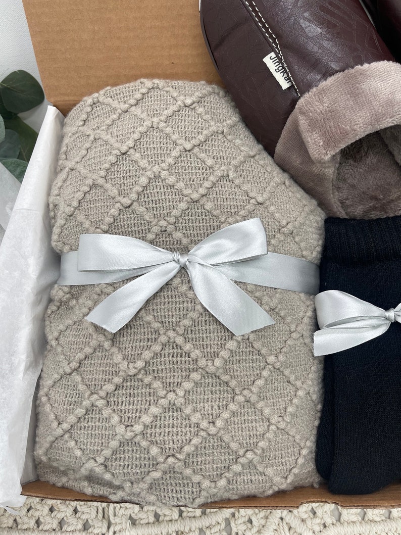 Pode incluir: Uma caixa de presente cheia de itens aconchegantes, incluindo uma manta de malha bege com uma fita prateada, um par de meias pretas e um par de pantufas marrons com uma etiqueta "JingKea".
