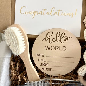 May include: A gift box filled with brown crinkle paper and a wooden sign that says "hello world" with spaces for the baby's date, time, length, and weight. The box also contains a white baby brush, a stainless steel straw, and a white and brown knitted toy.