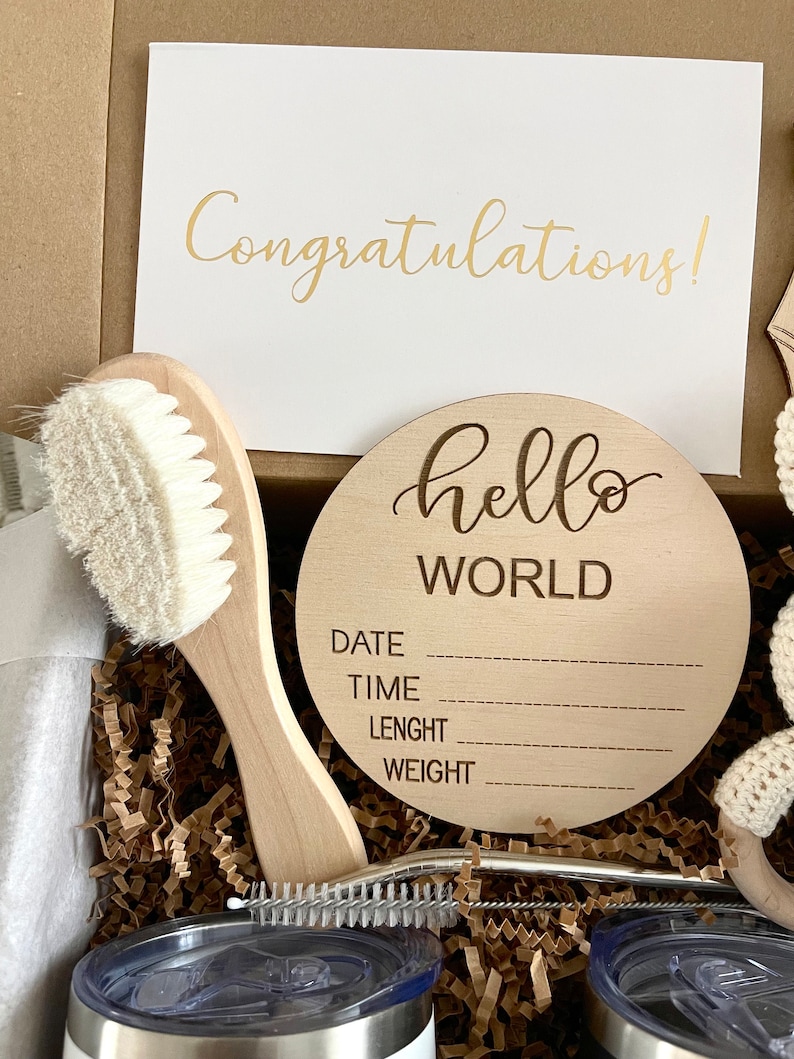 May include: A gift box filled with brown crinkle paper and a wooden sign that says "hello world" with spaces for the baby's date, time, length, and weight. The box also contains a white baby brush, a stainless steel straw, and a white and brown knitted toy.
