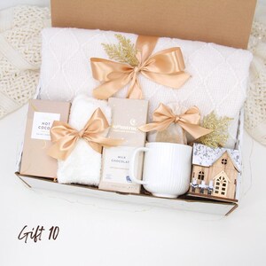 Puede incluir: Una caja de regalo llena de una manta blanca, chocolate caliente, chocolate con leche, una taza y una casa de madera decorativa. Los art&iacute;culos est&aacute;n atados con lazos dorados y colocados en una caja de cart&oacute;n. El texto "Gift 10" est&aacute; escrito en la parte inferior izquierda.