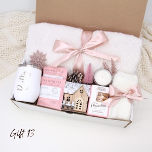 Puede incluir: Una caja de regalo llena de una manta blanca atada con una cinta rosa, un vaso blanco, paquetes de t&eacute;, una casa de madera, una vela, galletas y una peque&ntilde;a toalla blanca atada con una cinta rosa. La caja est&aacute; etiquetada como "Gift 13".