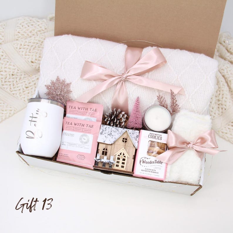 Puede incluir: Una caja de regalo llena de una manta blanca atada con una cinta rosa, un vaso blanco, paquetes de t&eacute;, una casa de madera, una vela, galletas y una peque&ntilde;a toalla blanca atada con una cinta rosa. La caja est&aacute; etiquetada como "Gift 13".
