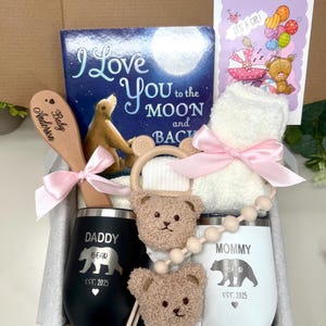 Może przedstawiać: Pudełko prezentowe dla dziecka z książką dla dzieci, drewnianą łyżką z napisem "Baby Anderson", białym ręcznikiem kąpielowym zawiązanym r&oacute;żową wstążką, gryzakiem, dwoma izolowanymi kubkami z napisami "Daddy Bear" i "Mommy Bear" oraz kartką z napisem "It's a Girl!"