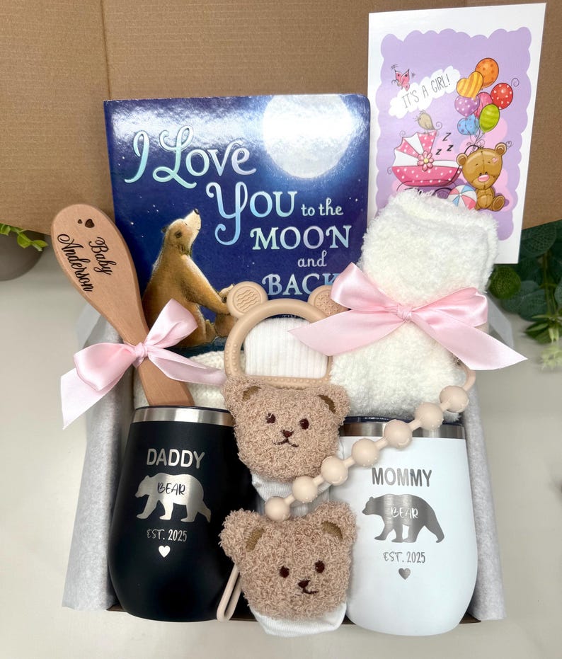 Może przedstawiać: Pudełko prezentowe dla dziecka z książką dla dzieci, drewnianą łyżką z napisem "Baby Anderson", białym ręcznikiem kąpielowym zawiązanym r&oacute;żową wstążką, gryzakiem, dwoma izolowanymi kubkami z napisami "Daddy Bear" i "Mommy Bear" oraz kartką z napisem "It's a Girl!"