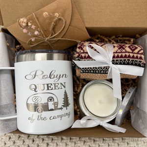 May include: A gift box filled with camping themed items. The box contains a white travel mug with the text "Robyn Queen of the camping" and a camper van graphic, a pair of brown and red patterned socks, a white candle in a glass jar, and a small roll of white ribbon.