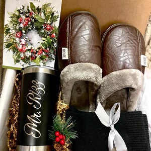 Pu&ograve; includere: Una scatola regalo piena di una variet&agrave; di articoli, tra cui un bicchiere termico nero e argento con il testo "Mr. Riggs" inciso su di esso, un paio di pantofole in pelle marrone con fodera in pelliccia sintetica, una sciarpa di lana nera con un nastro di raso bianco e un biglietto di auguri di Natale con un design a corona. La scatola &egrave; rivestita con carta triturata marrone.