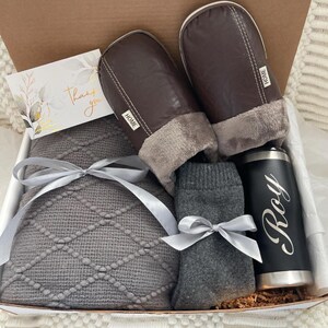 Pu&ograve; includere: Una scatola regalo piena di articoli accoglienti per una serata rilassante. Include una coperta di maglia grigia, un paio di pantofole in similpelle marrone con fodera in pile grigia, un paio di calzini grigi e un bicchiere in acciaio inossidabile nero con il nome "Roy" inciso.