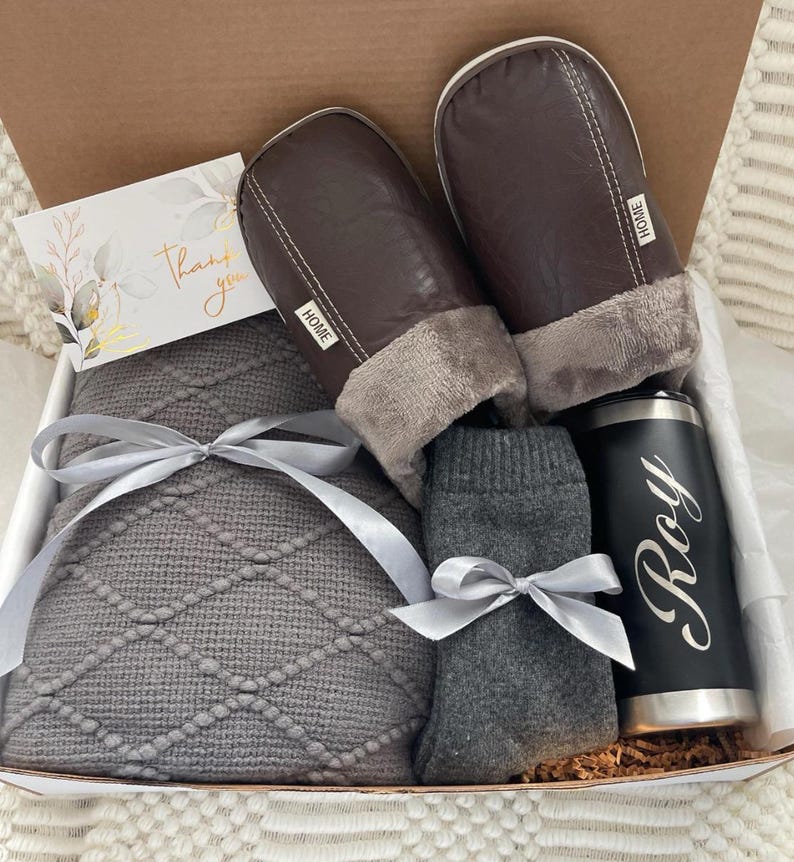 Pu&ograve; includere: Una scatola regalo piena di articoli accoglienti per una serata rilassante. Include una coperta di maglia grigia, un paio di pantofole in similpelle marrone con fodera in pile grigia, un paio di calzini grigi e un bicchiere in acciaio inossidabile nero con il nome "Roy" inciso.