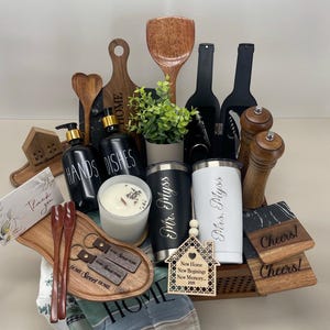 K&ouml;nnte beinhalten: Ein Geschenkkorb gef&uuml;llt mit Einweihungsgeschenken, darunter ein Holzbrett, ein Holzl&ouml;ffel, eine Kerze, zwei personalisierte Becher in Schwarz und Wei&szlig;, ein Holzhaus-Anh&auml;nger und zwei Untersetzer. Der Korb ist mit einem blau-wei&szlig; gestreiften Handtuch ausgelegt.