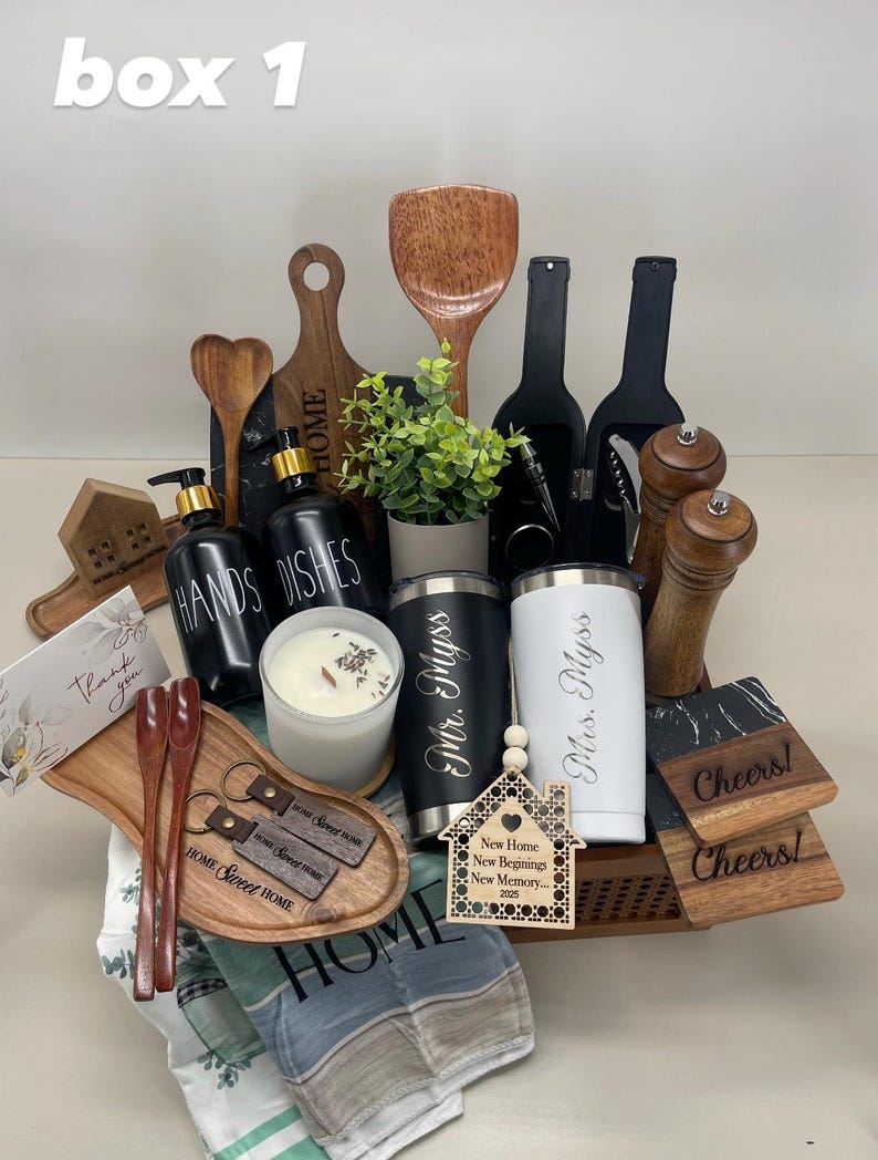K&ouml;nnte beinhalten: Ein Geschenkkorb gef&uuml;llt mit Einweihungsgeschenken, darunter ein Holzbrett, ein Holzl&ouml;ffel, eine Kerze, zwei personalisierte Becher in Schwarz und Wei&szlig;, ein Holzhaus-Anh&auml;nger und zwei Untersetzer. Der Korb ist mit einem blau-wei&szlig; gestreiften Handtuch ausgelegt.