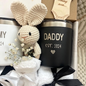 May include: A gift box filled with baby shower gifts for a new dad. The box includes a black and white mug that says "Daddy Est. 2024" with a small heart, a white mug that says "Mommy 2024", a crocheted bunny rattle, a pair of white baby socks, and a wooden cutout of a onesie that says "Baby coming soon..."