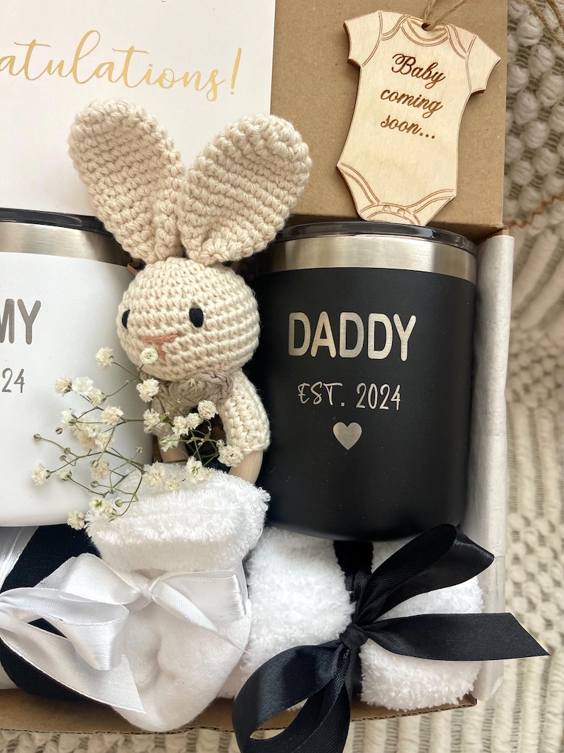 May include: A gift box filled with baby shower gifts for a new dad. The box includes a black and white mug that says "Daddy Est. 2024" with a small heart, a white mug that says "Mommy 2024", a crocheted bunny rattle, a pair of white baby socks, and a wooden cutout of a onesie that says "Baby coming soon..."