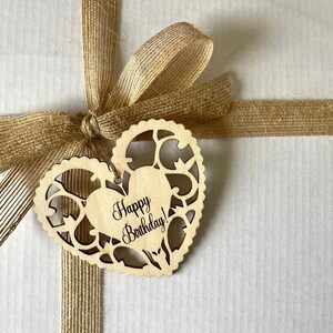 Pode incluir: Uma sacola de presente de papel castanho com uma fita de juta castanho amarrada em um la&ccedil;o. Uma etiqueta de madeira em forma de cora&ccedil;&atilde;o com o texto "Happy Birthday!" est&aacute; presa &agrave; fita.