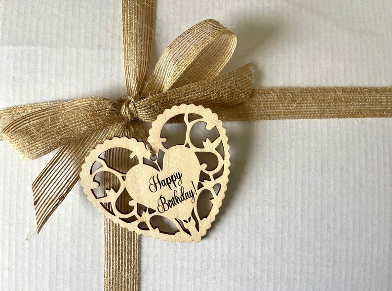 Pode incluir: Uma sacola de presente de papel castanho com uma fita de juta castanho amarrada em um la&ccedil;o. Uma etiqueta de madeira em forma de cora&ccedil;&atilde;o com o texto "Happy Birthday!" est&aacute; presa &agrave; fita.