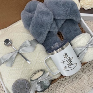 Op de afbeelding: Een witte cadeaumand gevuld met knusse items, waaronder een wit deken, een paar grijze fluffy pantoffels, een witte mok met de tekst "Drink Coffee", een witte kaars en een zilveren lepel.
