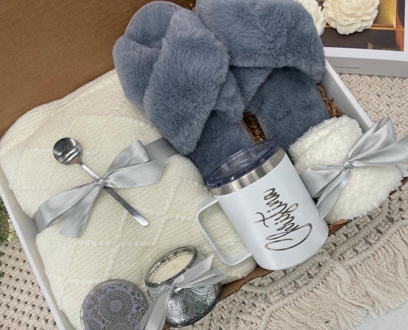 Op de afbeelding: Een witte cadeaumand gevuld met knusse items, waaronder een wit deken, een paar grijze fluffy pantoffels, een witte mok met de tekst "Drink Coffee", een witte kaars en een zilveren lepel.