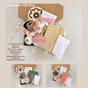 May include: An open gift box filled with self-care items. The box contains a candle, notebook, lip balm, tea, and a mirror with the text "You're awesome". The box also includes a wooden coaster and a silk hair tie. The text "Awesome Gift Box" is at the top.