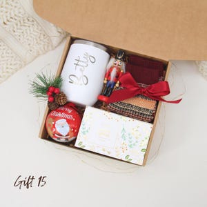 Puede incluir: Una caja de regalo navide&ntilde;a con un vaso blanco con "Betty" en dorado, un cascanueces, una bola roja con Pap&aacute; Noel, un lazo rojo y una caja de jab&oacute;n. La caja es marr&oacute;n y contiene calcetines burdeos.