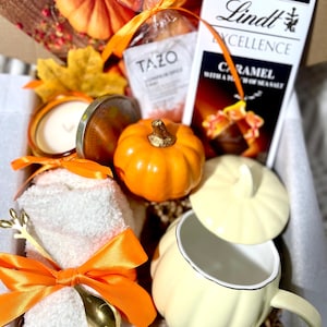 May include: A fall-themed gift box filled with items like a pumpkin-shaped mug, a candle, a towel, a spoon, a chocolate bar, and a tea bag. The box is decorated with fall leaves and pumpkins.