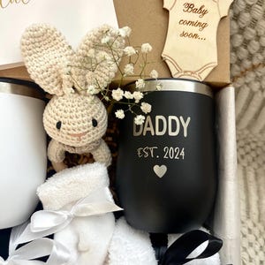 May include: A black and white gift box filled with a variety of items, including a black insulated tumbler with the text "Daddy Est. 2024", a white crocheted bunny, a pair of white baby socks, and a wooden baby onesie cutout with the text "Baby coming soon..."