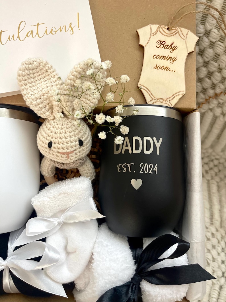 May include: A black and white gift box filled with a variety of items, including a black insulated tumbler with the text "Daddy Est. 2024", a white crocheted bunny, a pair of white baby socks, and a wooden baby onesie cutout with the text "Baby coming soon..."