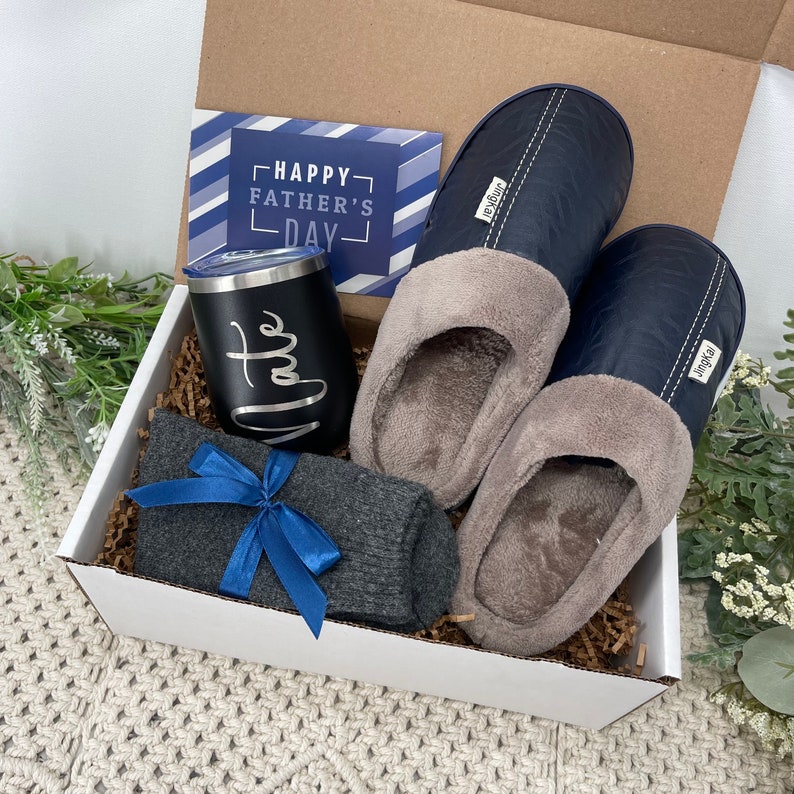 May include: A white gift box filled with a blue and gray patterned card that says "Happy Father's Day", a black insulated tumbler with the word "Date" written on it, a pair of blue and gray slippers, and a pair of gray socks with a blue ribbon.