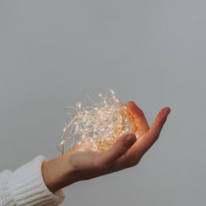 May include: A hand holding a string of white fairy lights, the lights are turned on and glowing.