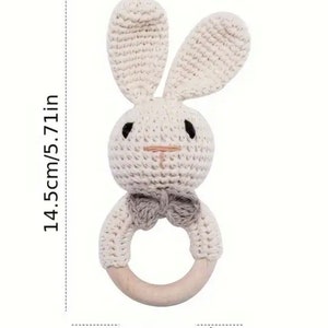 May include: A crocheted bunny rattle with a wooden ring. The bunny is cream coloured with a grey bow tie. The rattle measures 14.5cm tall and 5.8cm in diameter.