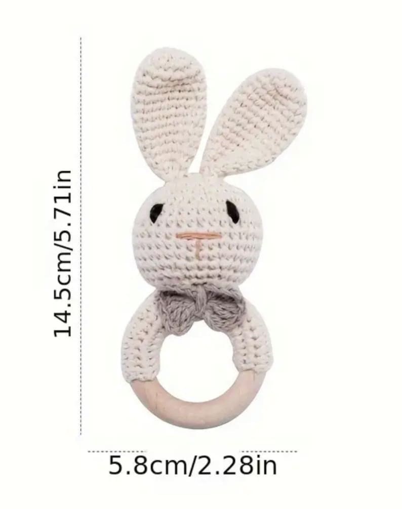 May include: A crocheted bunny rattle with a wooden ring. The bunny is cream coloured with a grey bow tie. The rattle measures 14.5cm tall and 5.8cm in diameter.