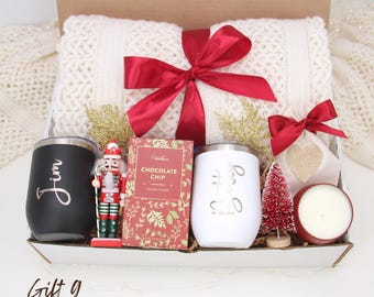 May include: A gift box with a cream-colored blanket tied with a red bow. It includes a black tumbler with "Jim" in cursive, a nutcracker, chocolate, a white tumbler, a small tree, a candle, and a small bag.