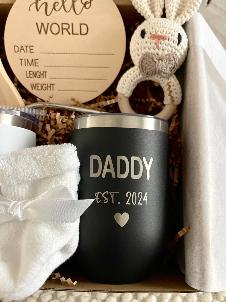 May include: A black stainless steel tumbler with the text "Daddy Est. 2024" and a small heart. The tumbler is in a gift box with other items, including a white towel, a wooden sign that says "hello world" and a crocheted bunny rattle.