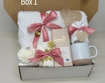 May include: A gift box with a white blanket, a mug, chocolate, and decorative items. The box is tied with pink ribbons and includes a gold heart-shaped ornament and a white flower. The text "Box 1" is visible.
