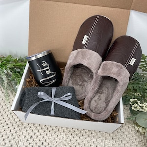 May include: A white gift box filled with a gray pair of socks, a black insulated tumbler with the word "Nate" on it, and a pair of brown leather slippers.